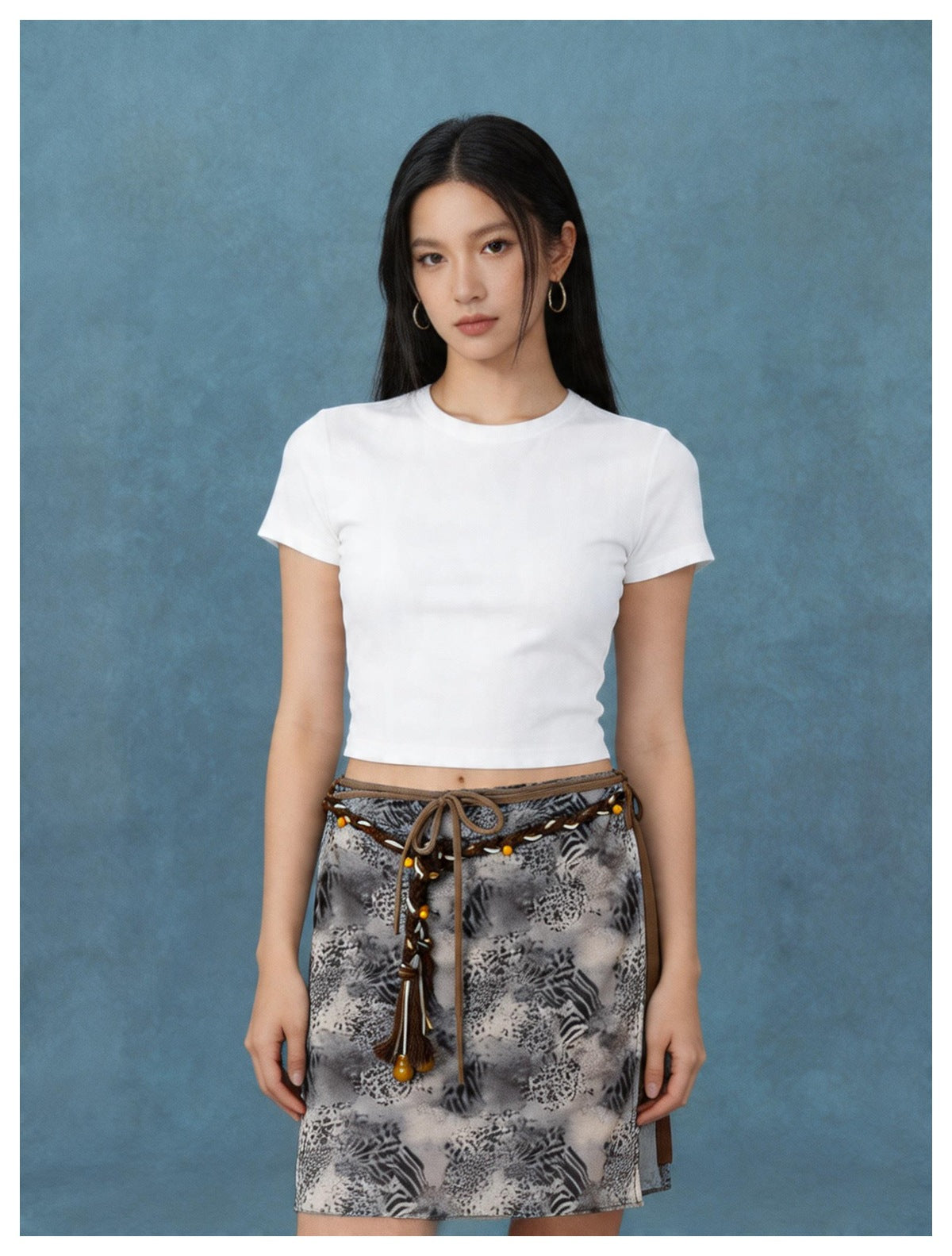 Model wearing an animal print wrap mini skirt with braided beaded cord belt and a white fitted tee against a blue backdrop