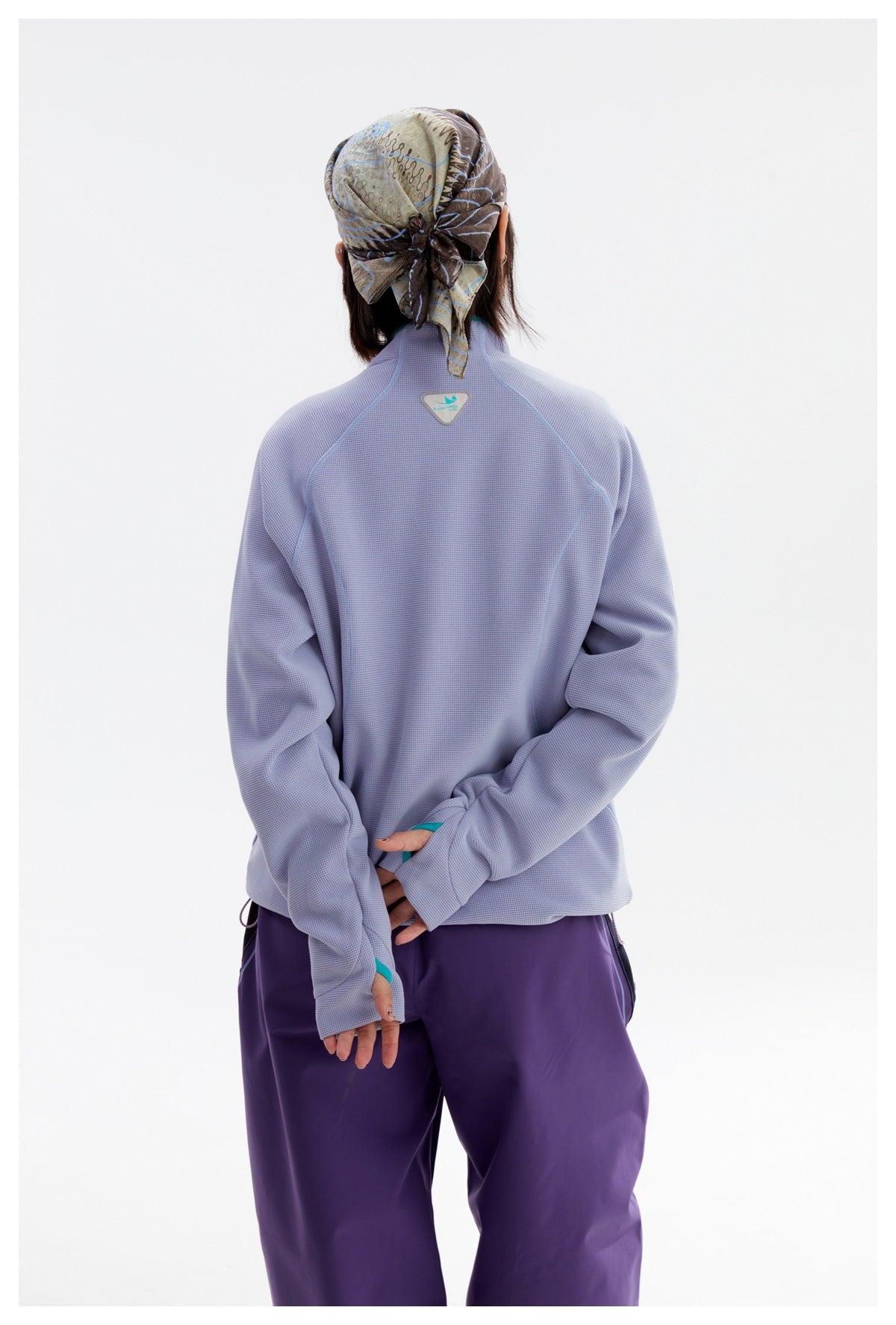 Back view of a model wearing a pale lavender jacket with teal seam details and loose purple pants.
