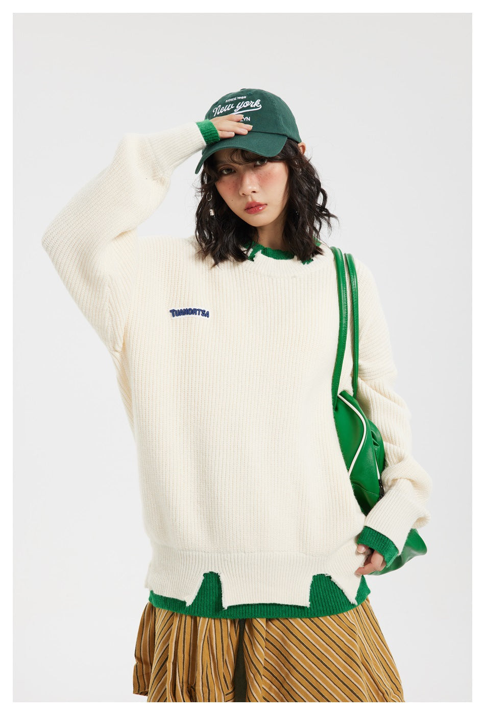 Close view of model in off-white ribbed sweater with green layered trim, blue patch, green cap and green bag