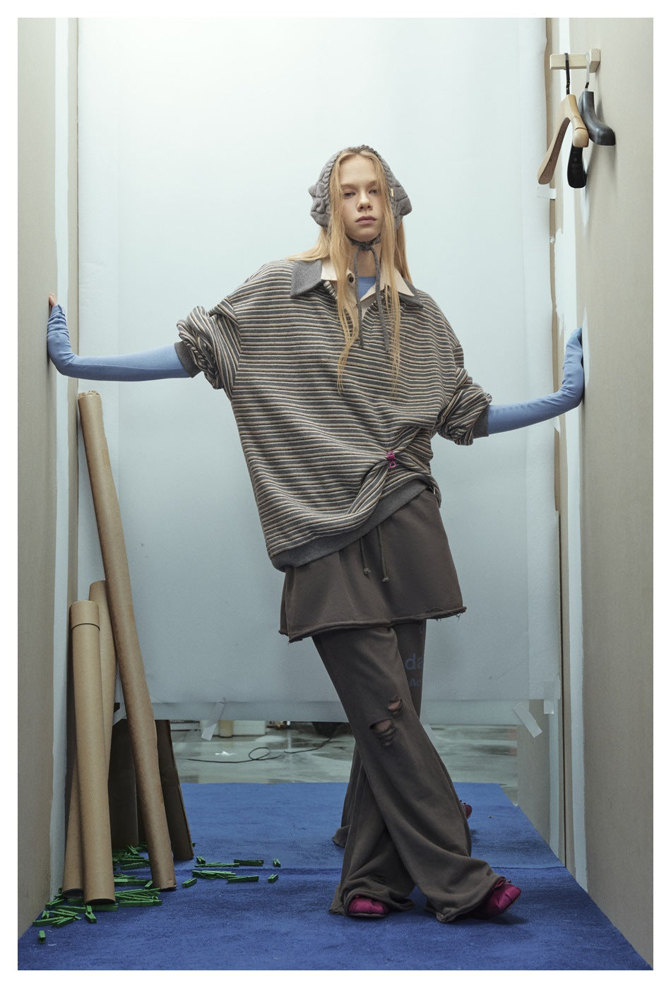 Model wearing a loose gray striped polo over blue sleeves and dark wide-leg pants, posing with arms outstretched.