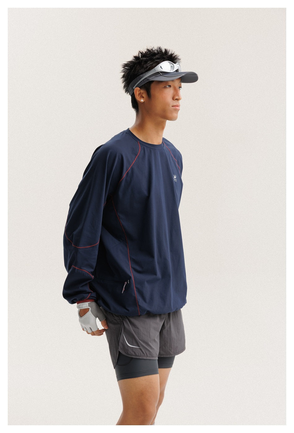 Model wearing a navy long-sleeve athletic top with red seam accents, gray shorts, gloves and a visor, shown from the front.