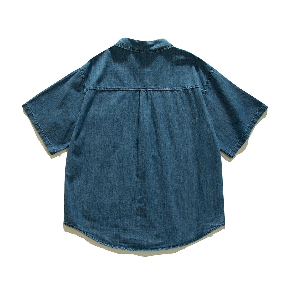 Back view of a short-sleeve blue denim shirt laid flat, with a center pleat and curved hem.