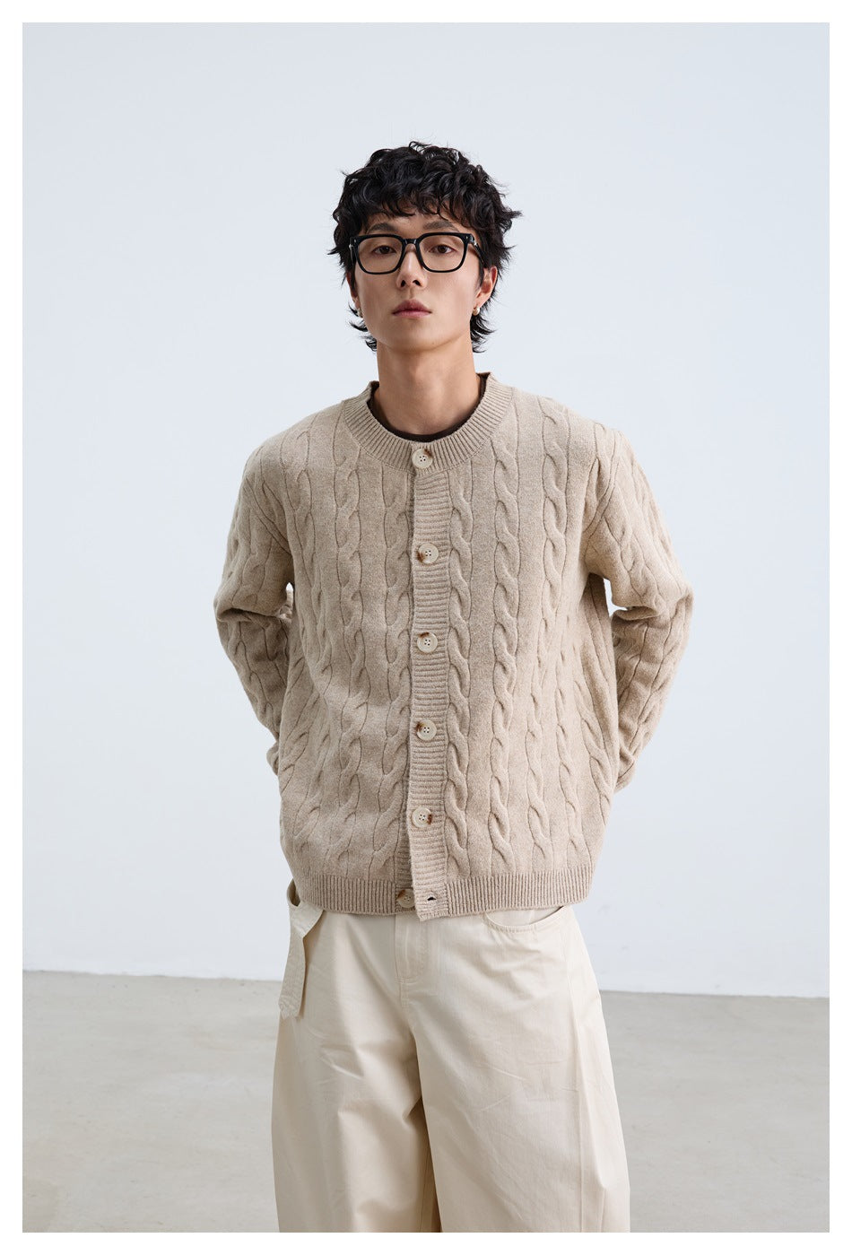 Model wearing a light beige cable-knit button cardigan and loose white trousers, facing forward.