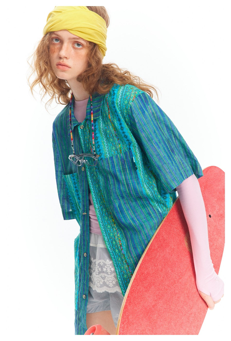 Model in a blue-green patterned button shirt over pink sleeves, holding a red skateboard.