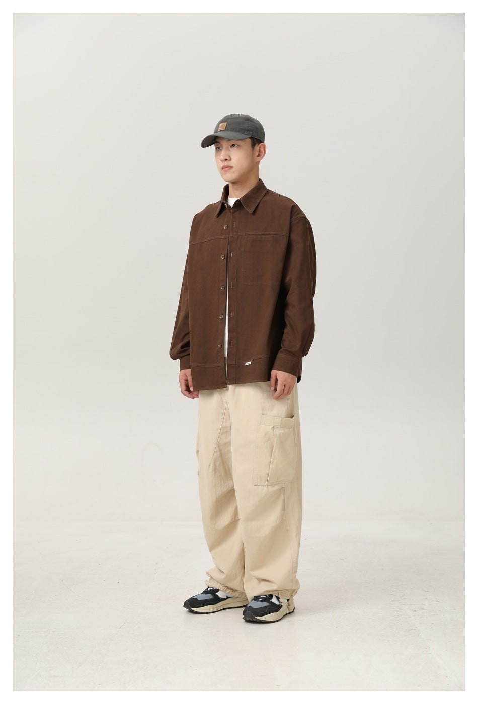 Man in a brown button-up shirt, white tee, beige cargo pants and a gray cap, shown in three-quarter view