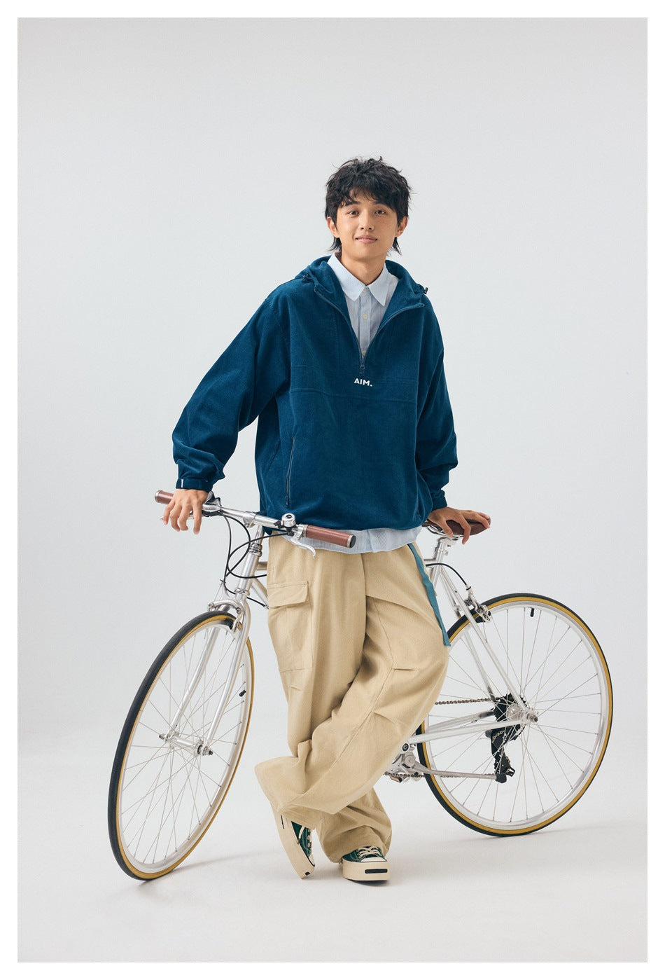 Model standing with a bicycle in a deep teal hooded pullover with small white AIM lettering.