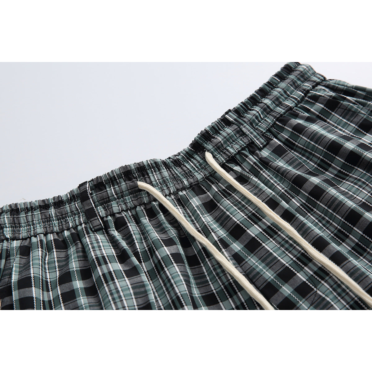 Close-up of black, gray, and muted green plaid shorts with an elastic waistband and cream drawstrings.