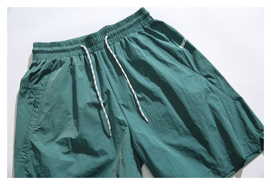 Teal flat-lay shorts with a gathered waistband, long white drawstrings, a zip pocket, and partial white lettering.