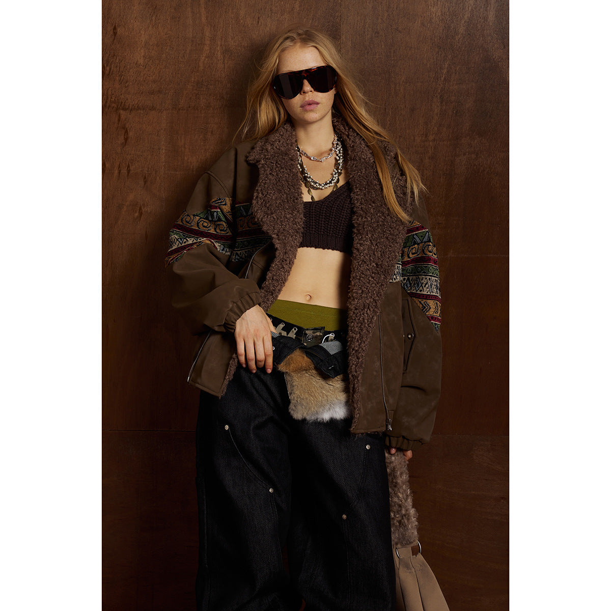 Model wearing a brown shearling-collar jacket with colorful patterned bands, black crop top and wide black jeans.