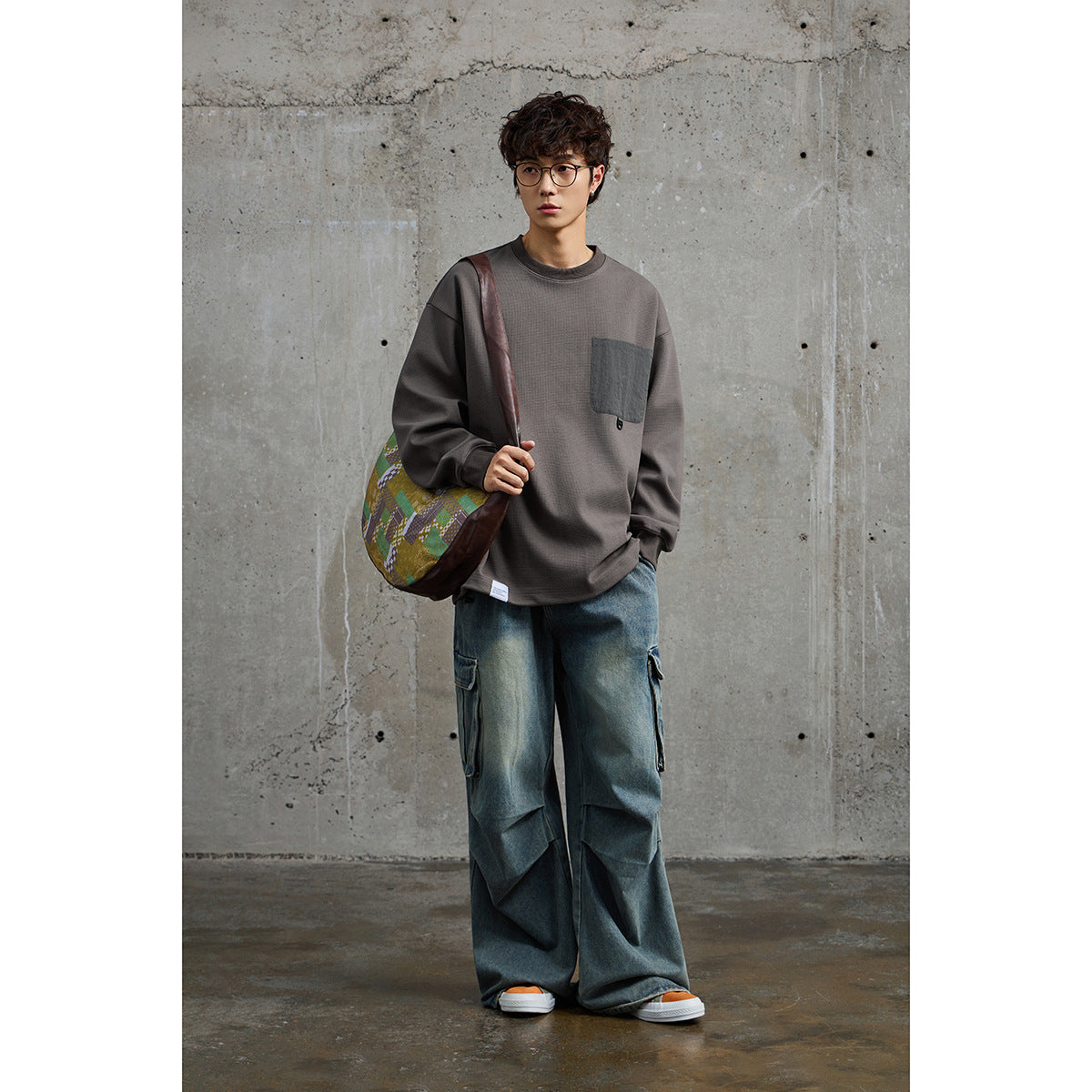 Model wearing a taupe crewneck sweatshirt with a dark chest pocket and loose blue cargo jeans.
