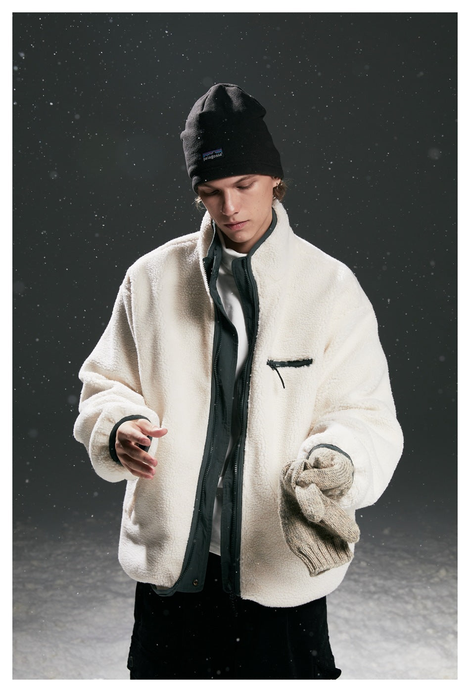 Model wearing a cream fleece zip jacket with dark trim and chest pocket, holding a knit glove against a snowy backdrop.