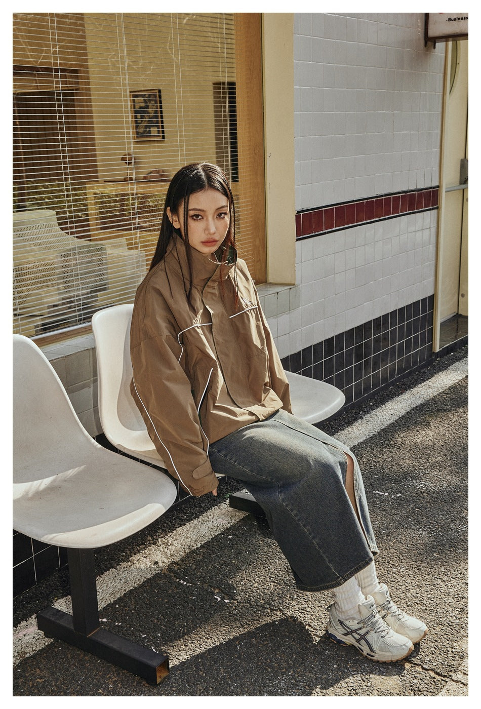 A model sits outdoors wearing a brown high-collar jacket with white piping and green accents, a long denim skirt, and