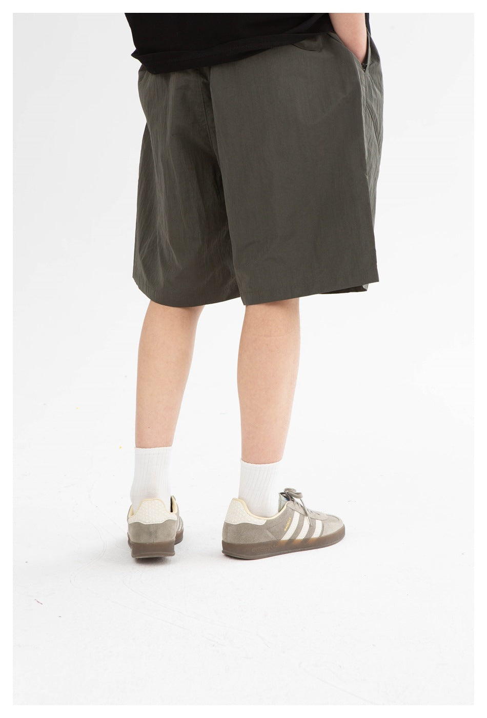 Rear three-quarter view of a model wearing dark olive knee-length shorts, white socks and light sneakers.