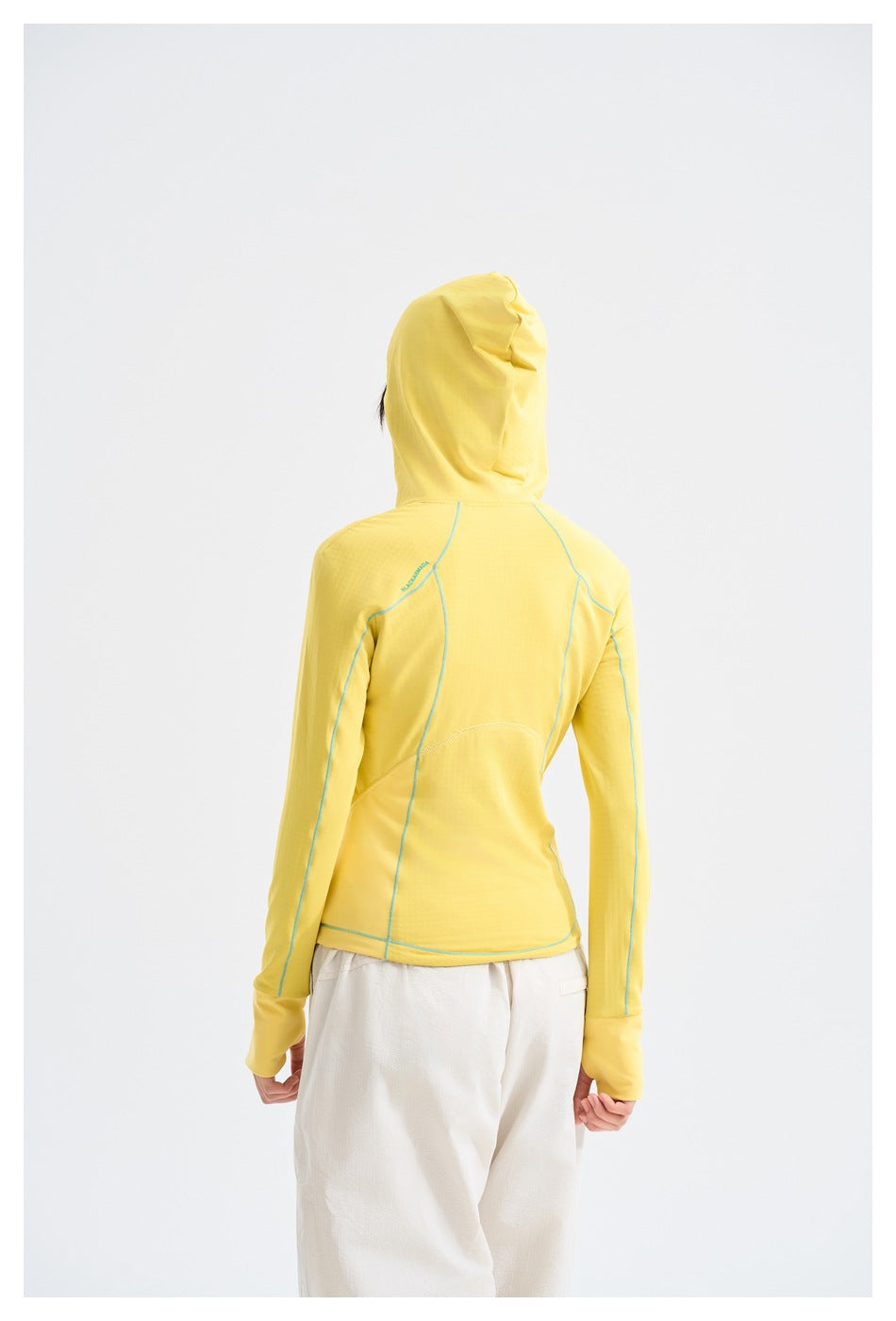 Back view of a model wearing a yellow hooded jacket with turquoise seam details and white pants.