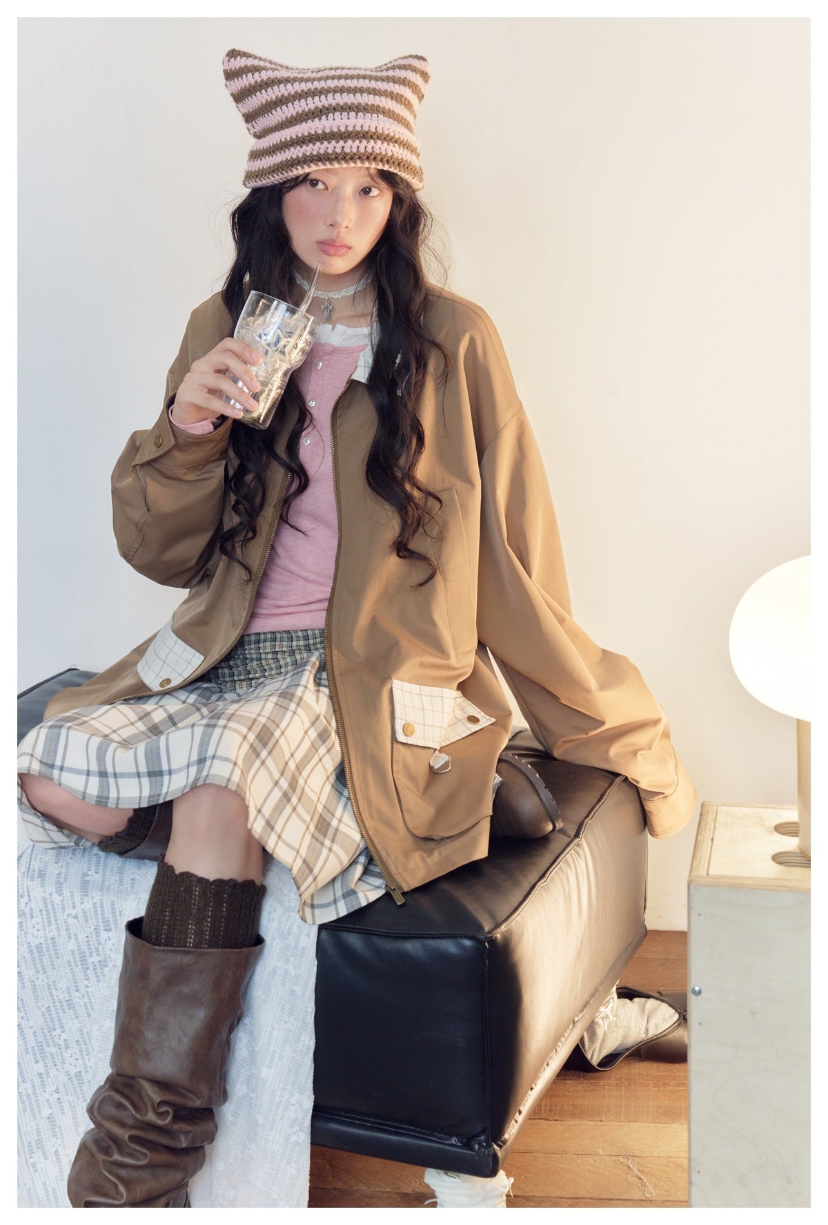 Seated model wearing an open tan jacket with plaid details over a pink top and plaid skirt; striped knit cat-ear hat.