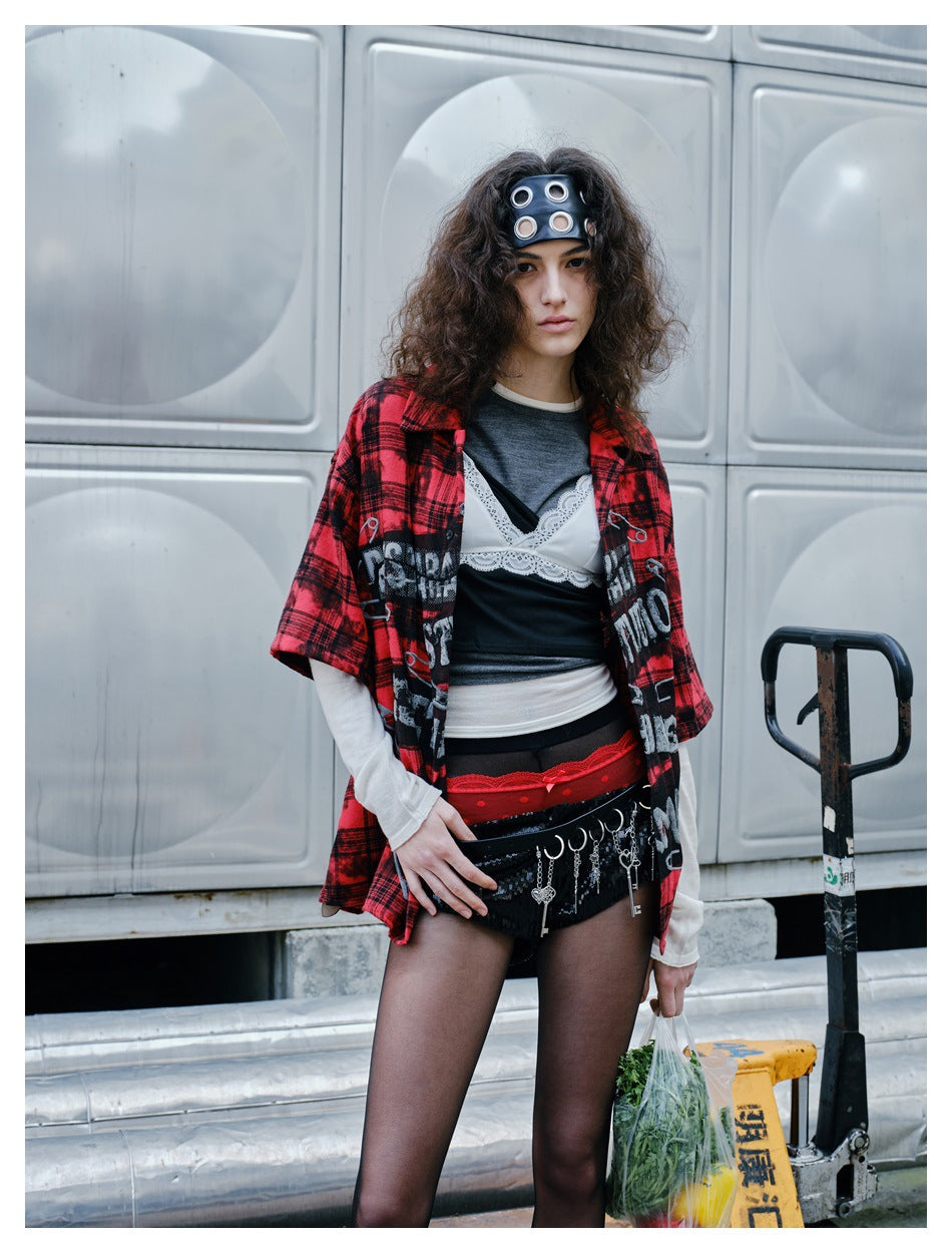 Model wearing a red-and-black plaid short-sleeve shirt with a safety-pin graphic over layered tops.