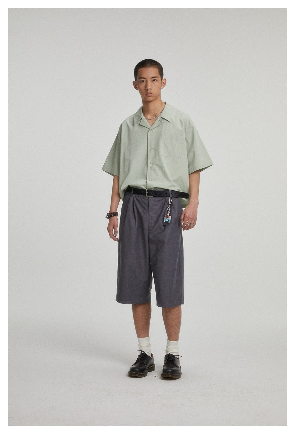 Model wearing gray pleated knee-length shorts with a green short-sleeve shirt, black belt and black shoes