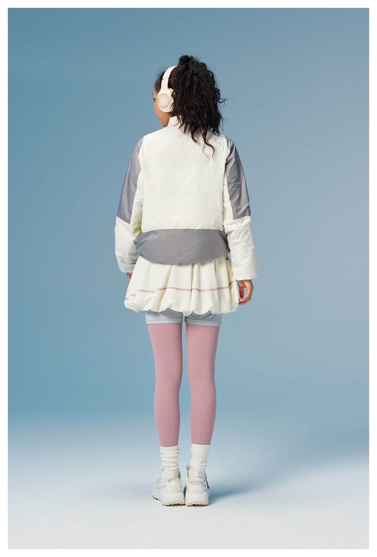 Rear view of a woman wearing a white jacket with gray panels and a white skirt over gray shorts and pink leggings.