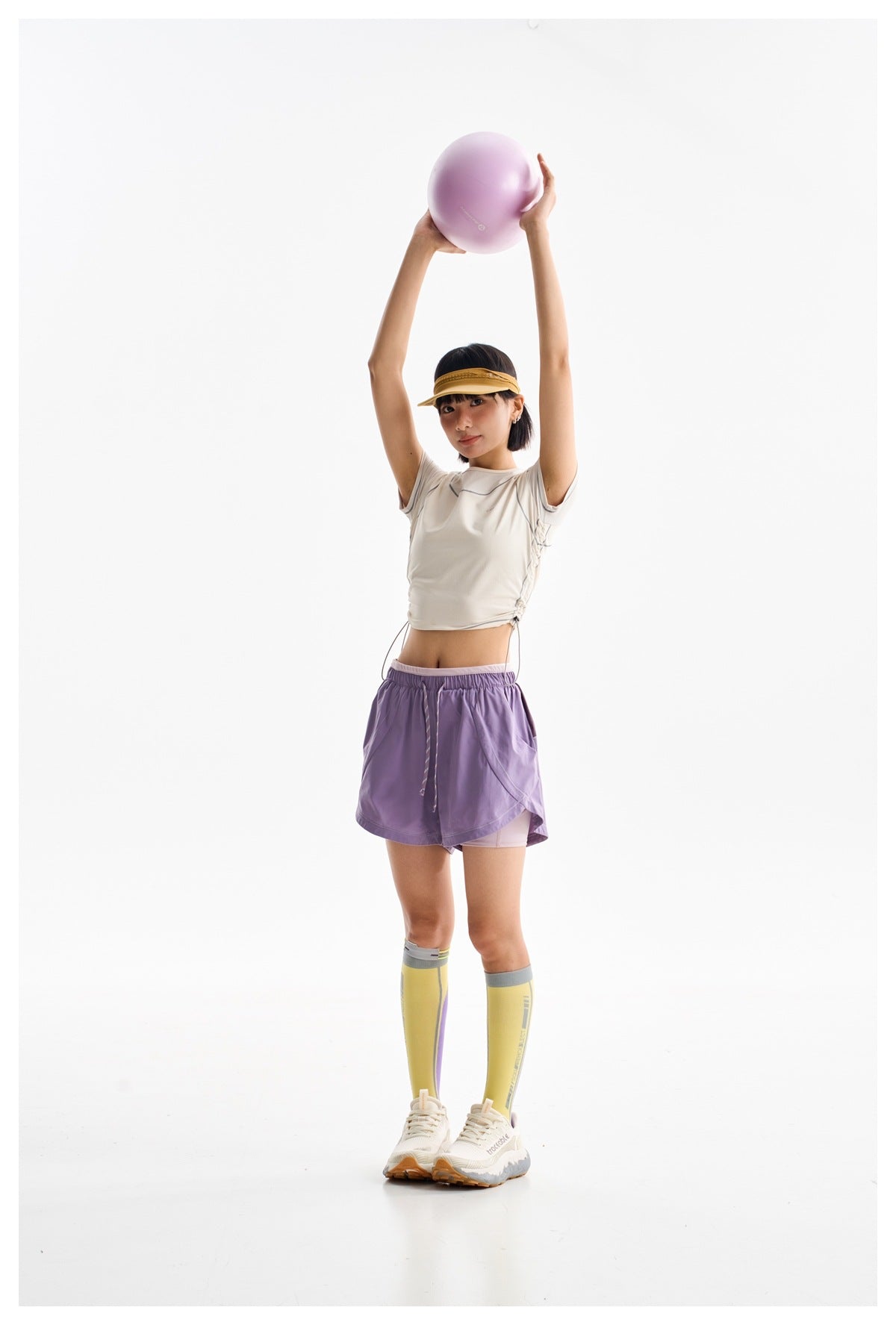 Model in a white contrast-seam athletic tee and lavender shorts, holding a lavender ball overhead.