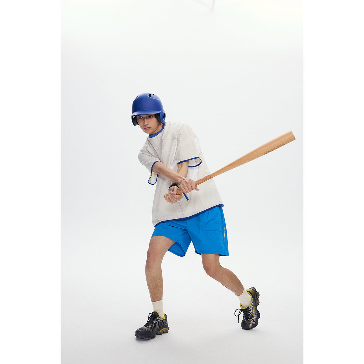 Model in an ivory shirt with blue trim and blue shorts, swinging a baseball bat.