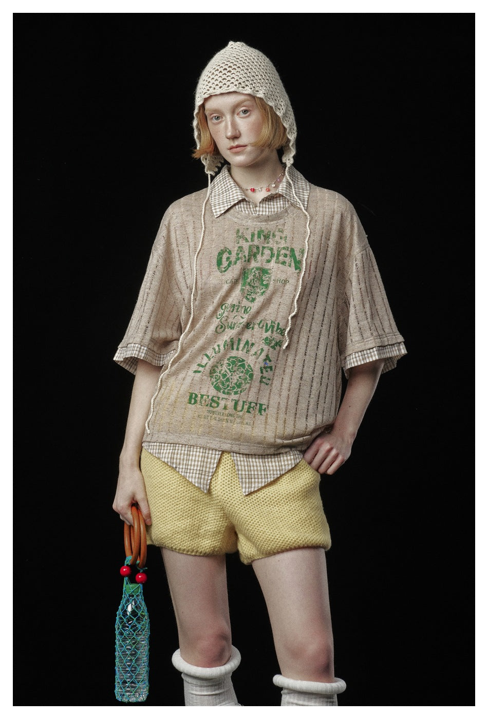 Model wearing a beige striped short-sleeve top with green graphic, checked shirt, yellow knit shorts and cream crocheted