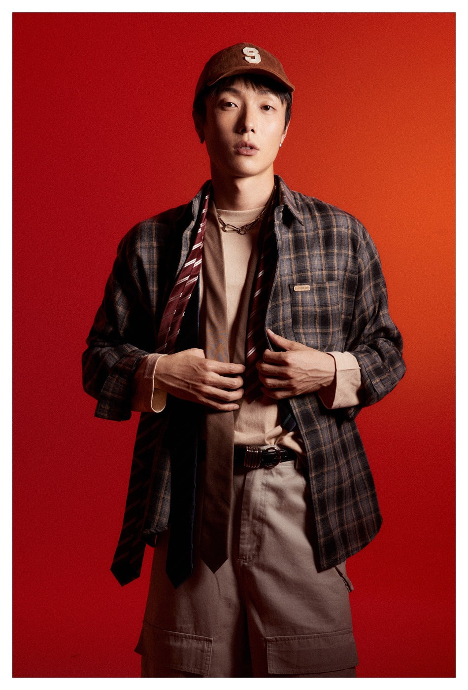 Front view of a model wearing a dark brown plaid button-up shirt over a beige top, with a brown cap and khaki shorts.
