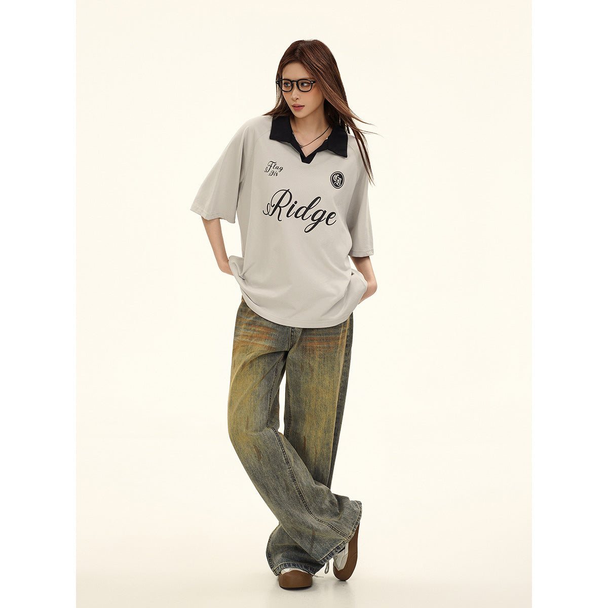 Model wearing an oversized light gray polo with black collar and lettering, paired with loose faded olive-gray jeans.
