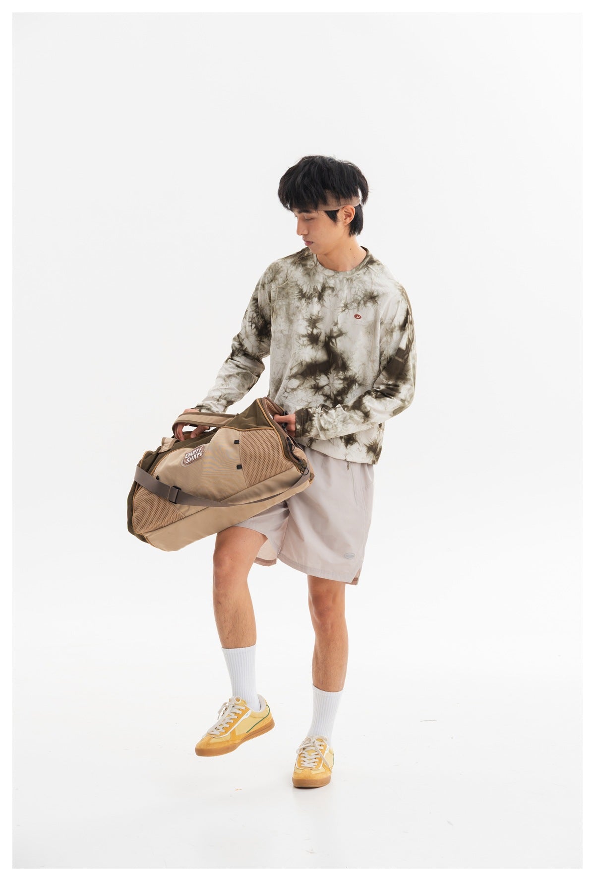 Model wearing an olive-gray tie-dye long-sleeve shirt and pale gray shorts while holding a tan duffel bag.