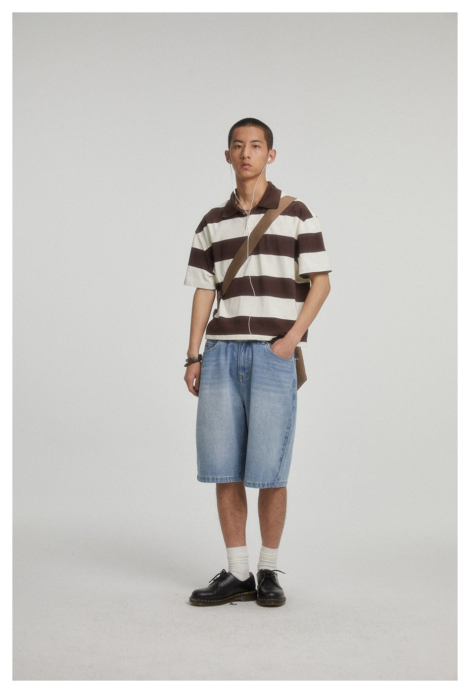 Man wearing light blue washed denim knee-length shorts with a brown and cream striped polo, crossbody strap and black shoes