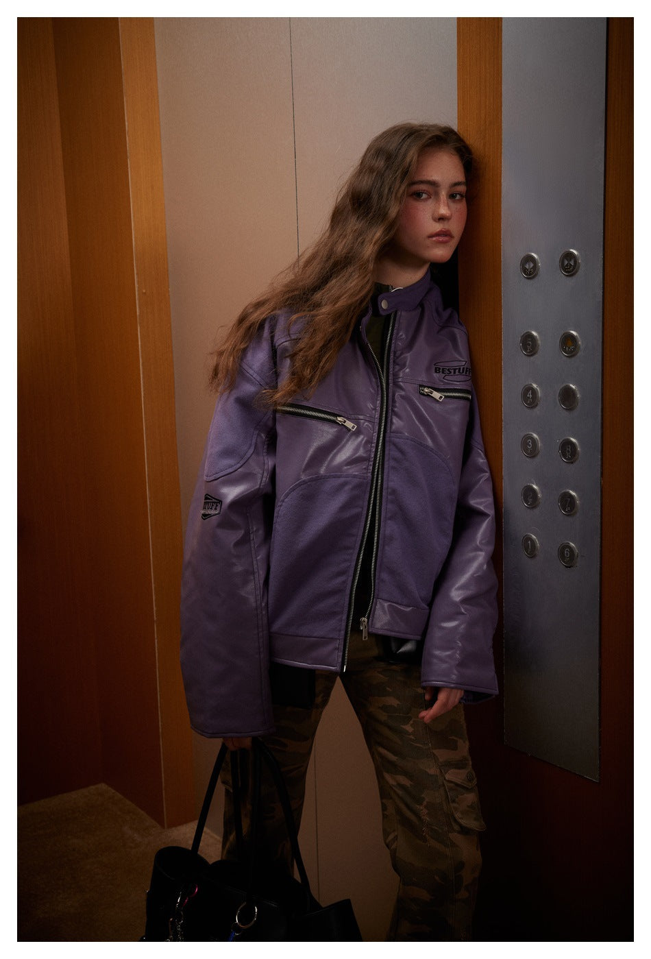 Model wearing a purple zip-front jacket with chest zippers and camouflage trousers beside an elevator panel.