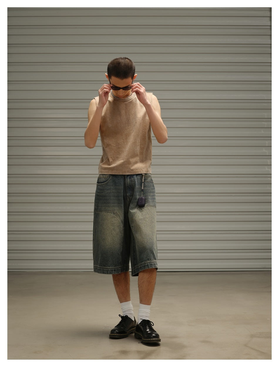 A man looks down while wearing a tan sleeveless top and loose, faded dark denim shorts.