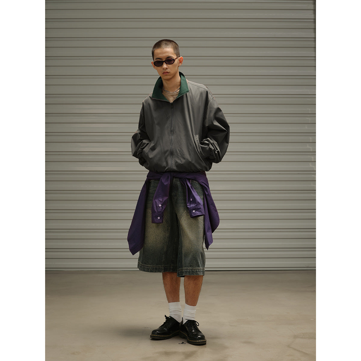 Model wearing a charcoal zip jacket with a dark green collar, oversized faded denim shorts, and a purple shirt tied at the