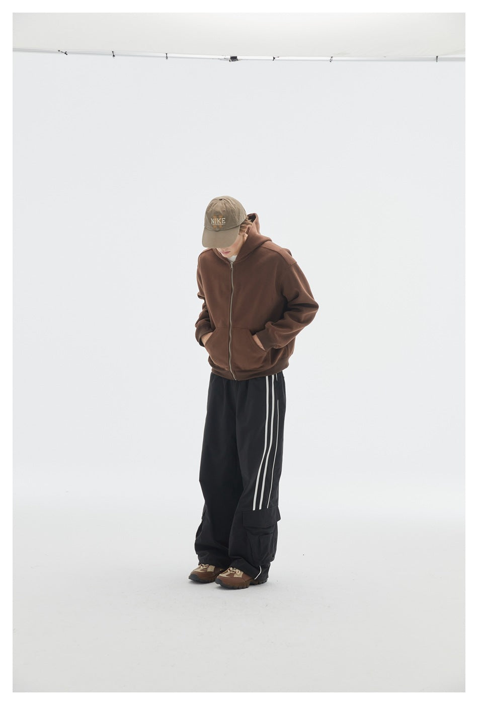 Model wearing a brown zip-up hoodie with front pockets and black pants with white side stripes.