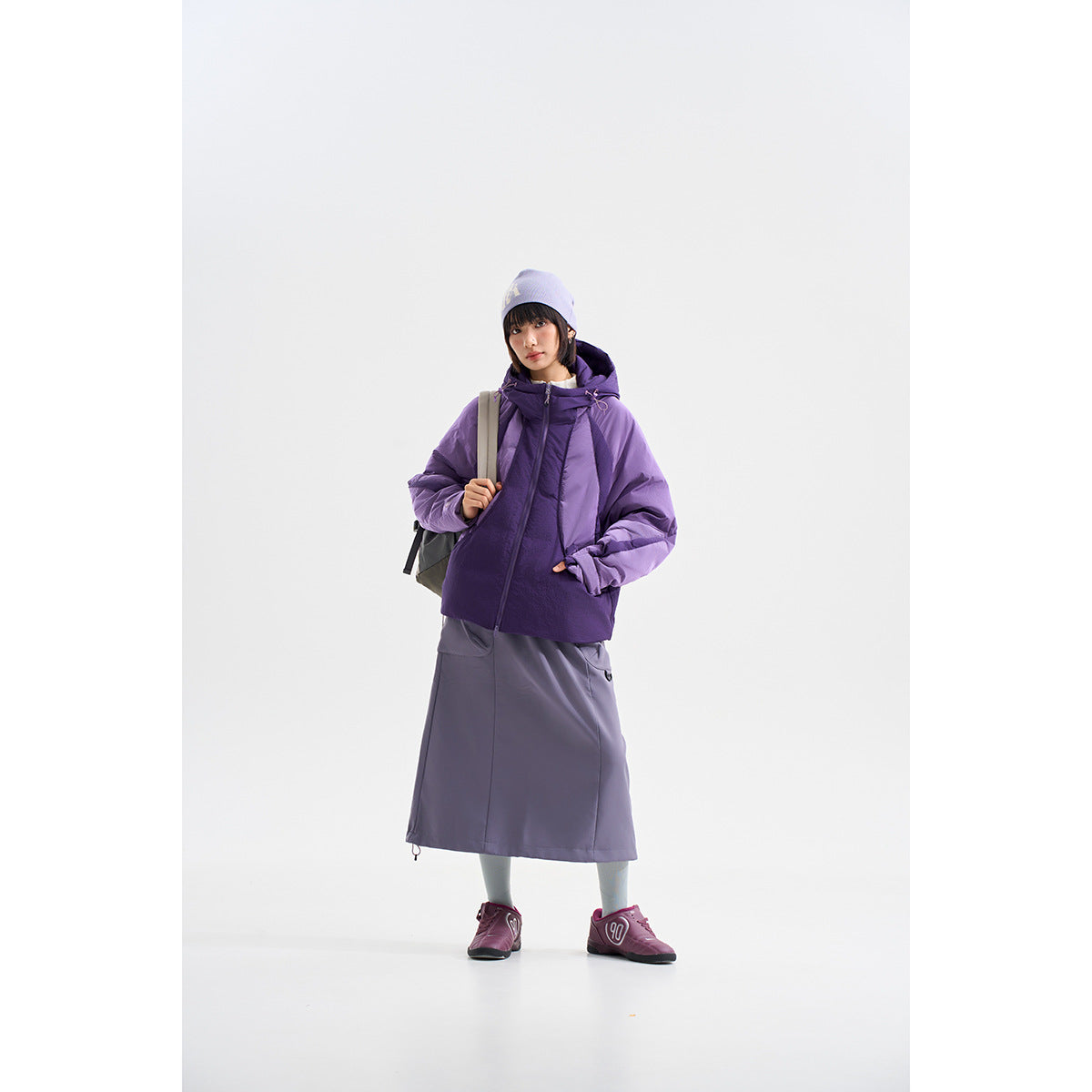 Model wears a two-tone purple hooded jacket with a long gray-purple skirt and a lavender beanie.