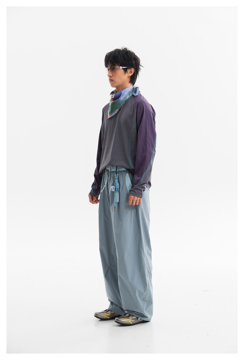 Full-length view of a model wearing a charcoal top with purple sleeves and teal seams, loose blue-gray trousers, and a