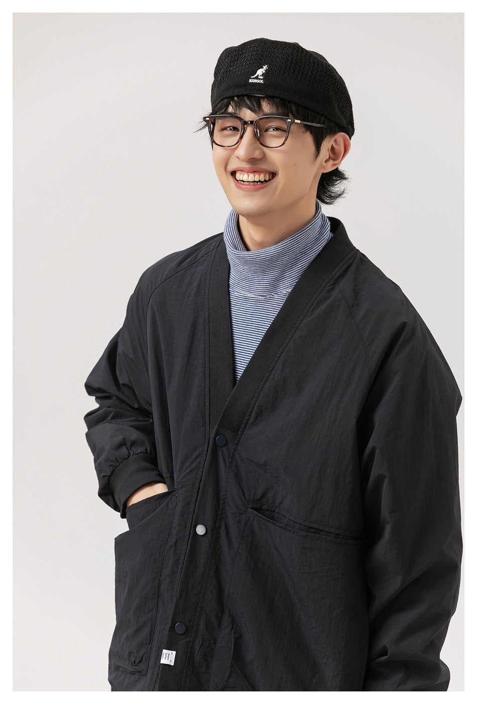 Model wearing a black snap-front jacket with large pockets over a striped turtleneck, shown from the waist up.
