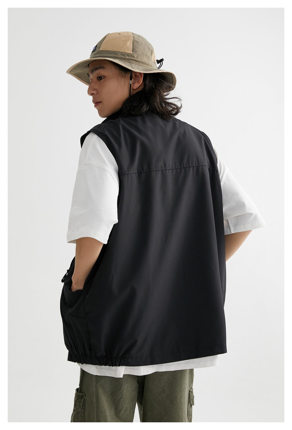 Rear view of a model wearing a black sleeveless vest over a white T-shirt, with a tan wide-brim hat and olive cargo pants.