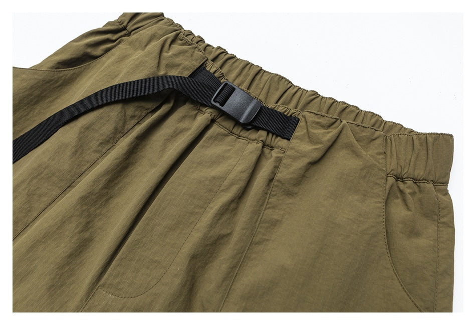 Close-up of olive pants showing an elastic waistband, black adjustable webbing strap, buckle, and pocket seams.