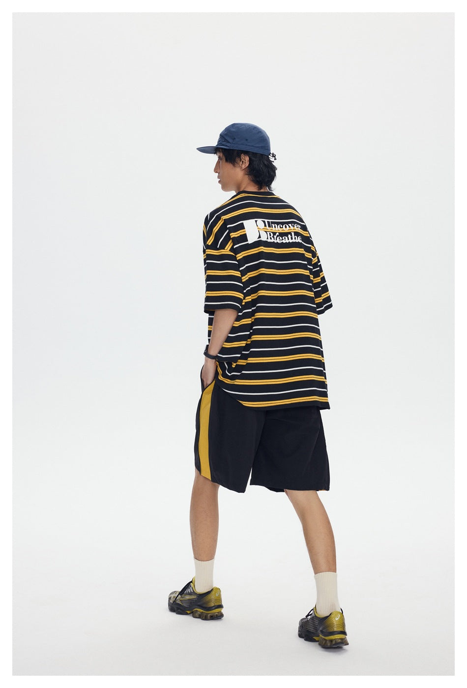 Model in black yellow-striped T-shirt, back three-quarter view, with black shorts and blue cap