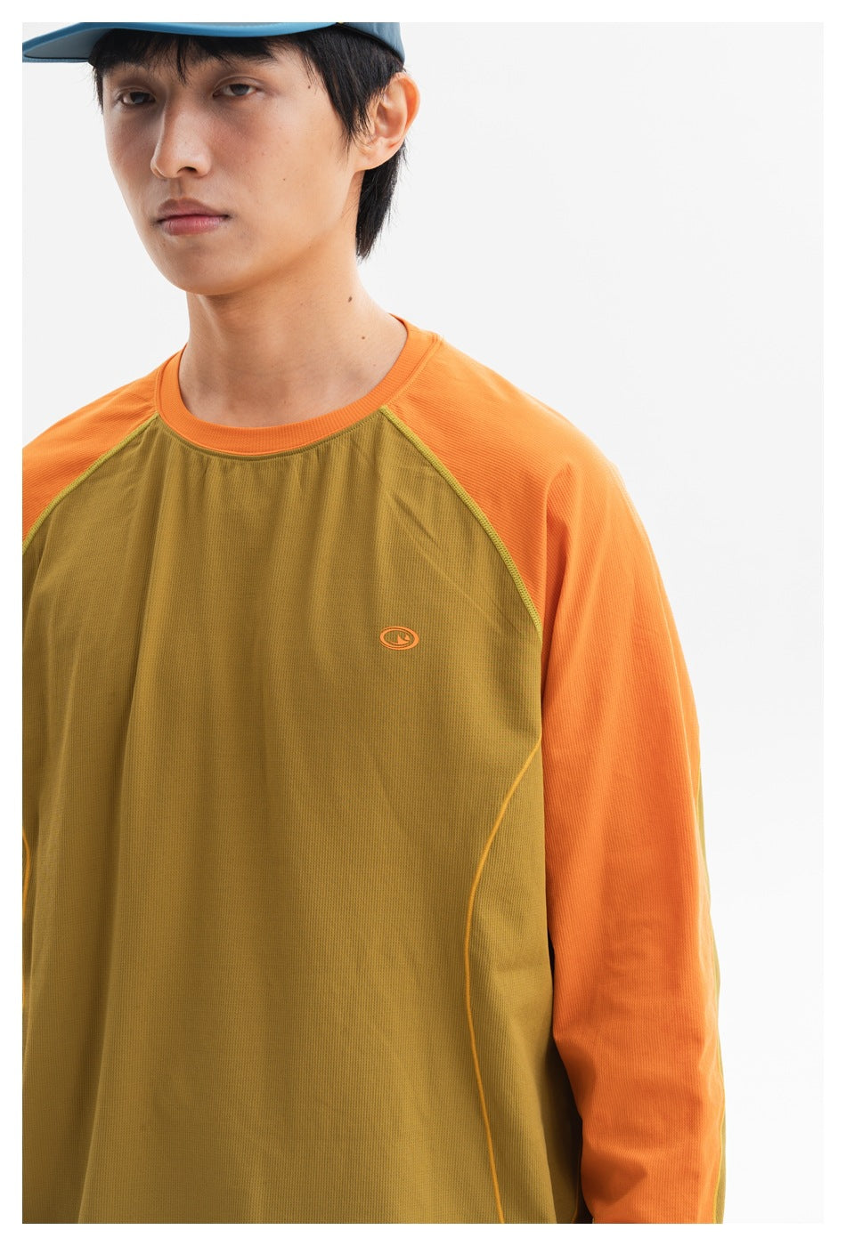 Close-up of a model wearing an olive-brown long-sleeve top with bright orange raglan sleeves and a small chest emblem.
