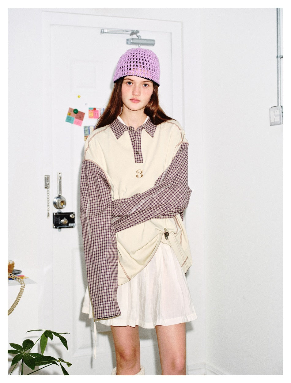 Front view of a model in an oversized cream top with brown plaid sleeves and collar, paired with a white pleated skirt.