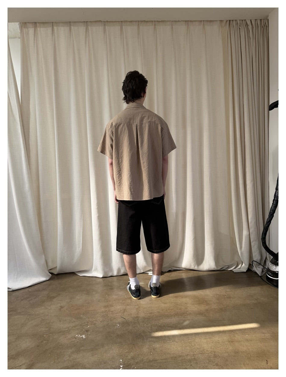 Back view of a man wearing khaki striped short-sleeve shirt with black denim shorts and sneakers