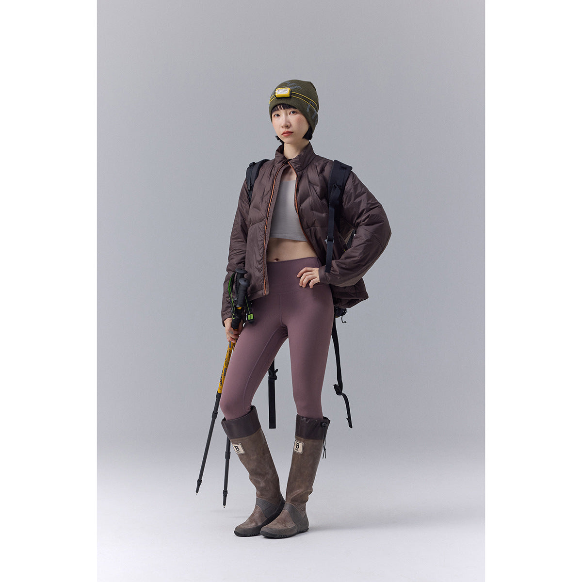 Model wears a dark brown quilted jacket, mauve leggings, tall brown boots, and an olive beanie with hiking gear.