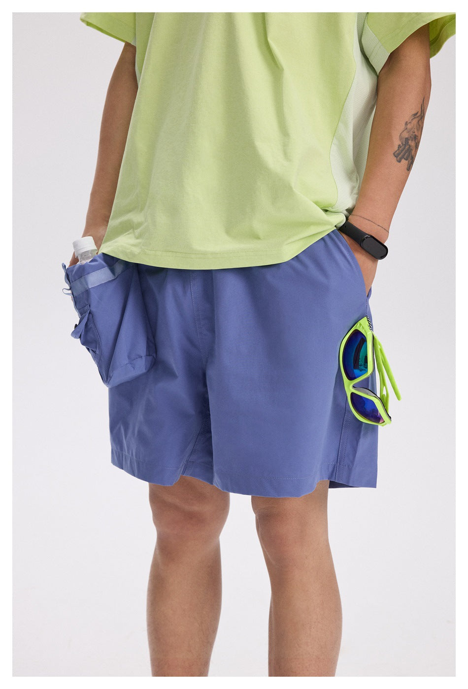 Cropped view of a person wearing blue-violet utility shorts with a lime-green shirt and sunglasses clipped to a pocket.