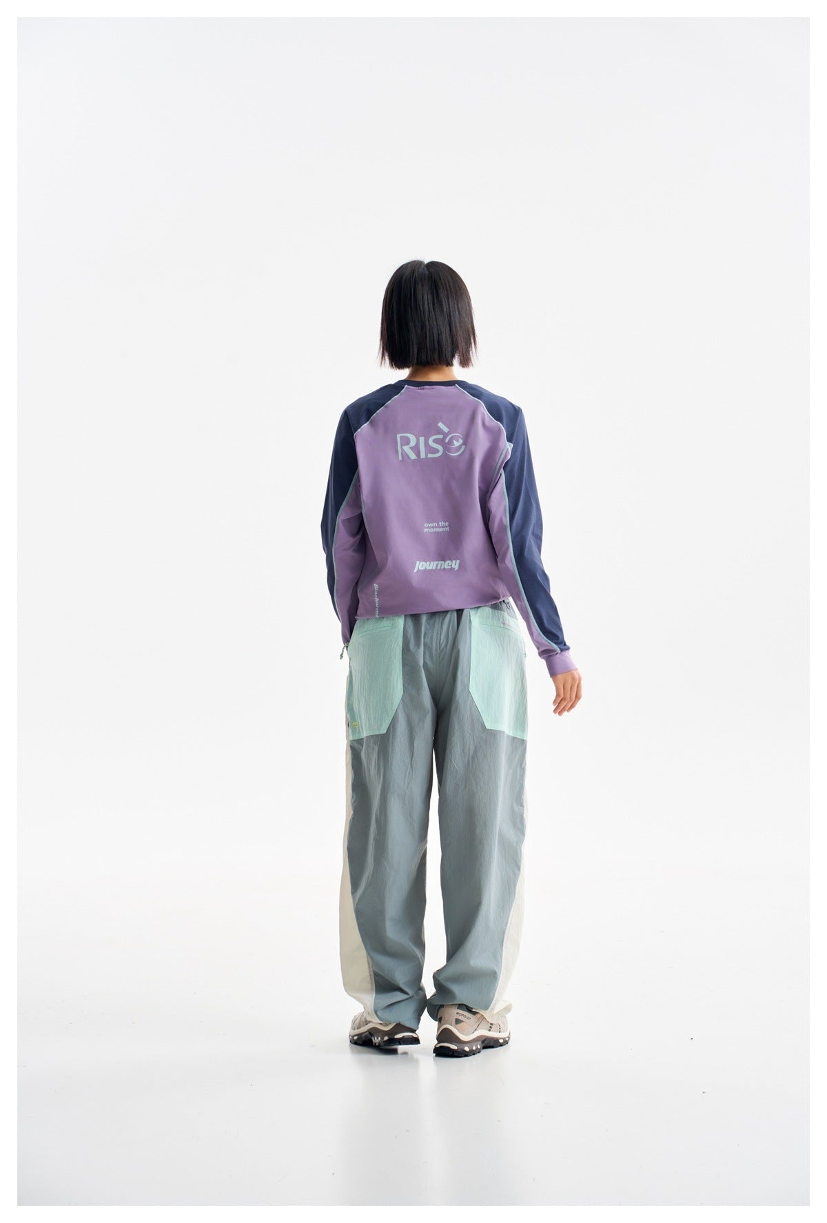 Rear view of model in a lavender top with navy sleeves and loose gray-green pants; the shirt has printed lettering.