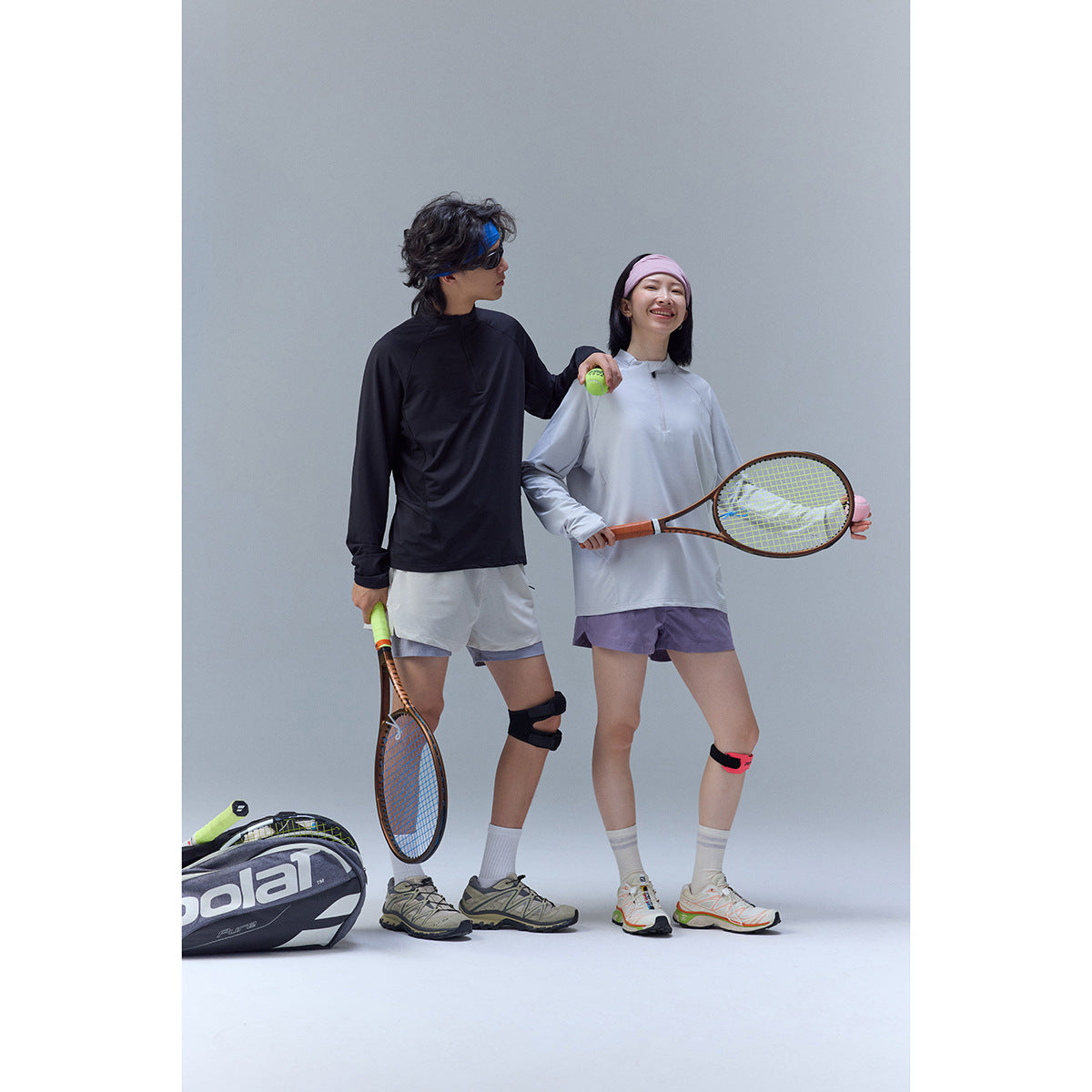 Two tennis players pose in black and pale gray long-sleeve tops with athletic shorts, holding rackets.