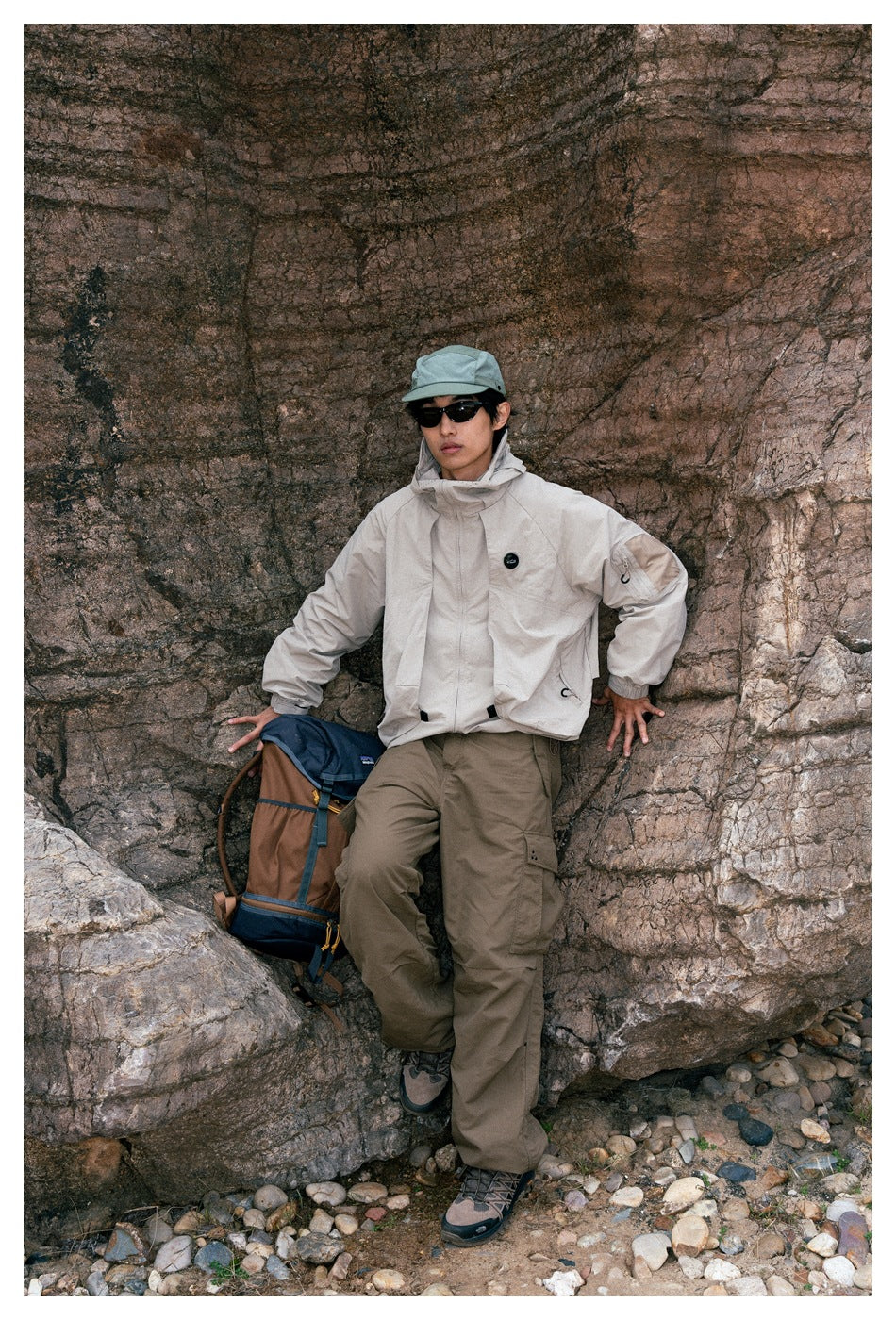 Person wearing a light beige hooded jacket and olive cargo pants, seated by a rock with a backpack beside them.