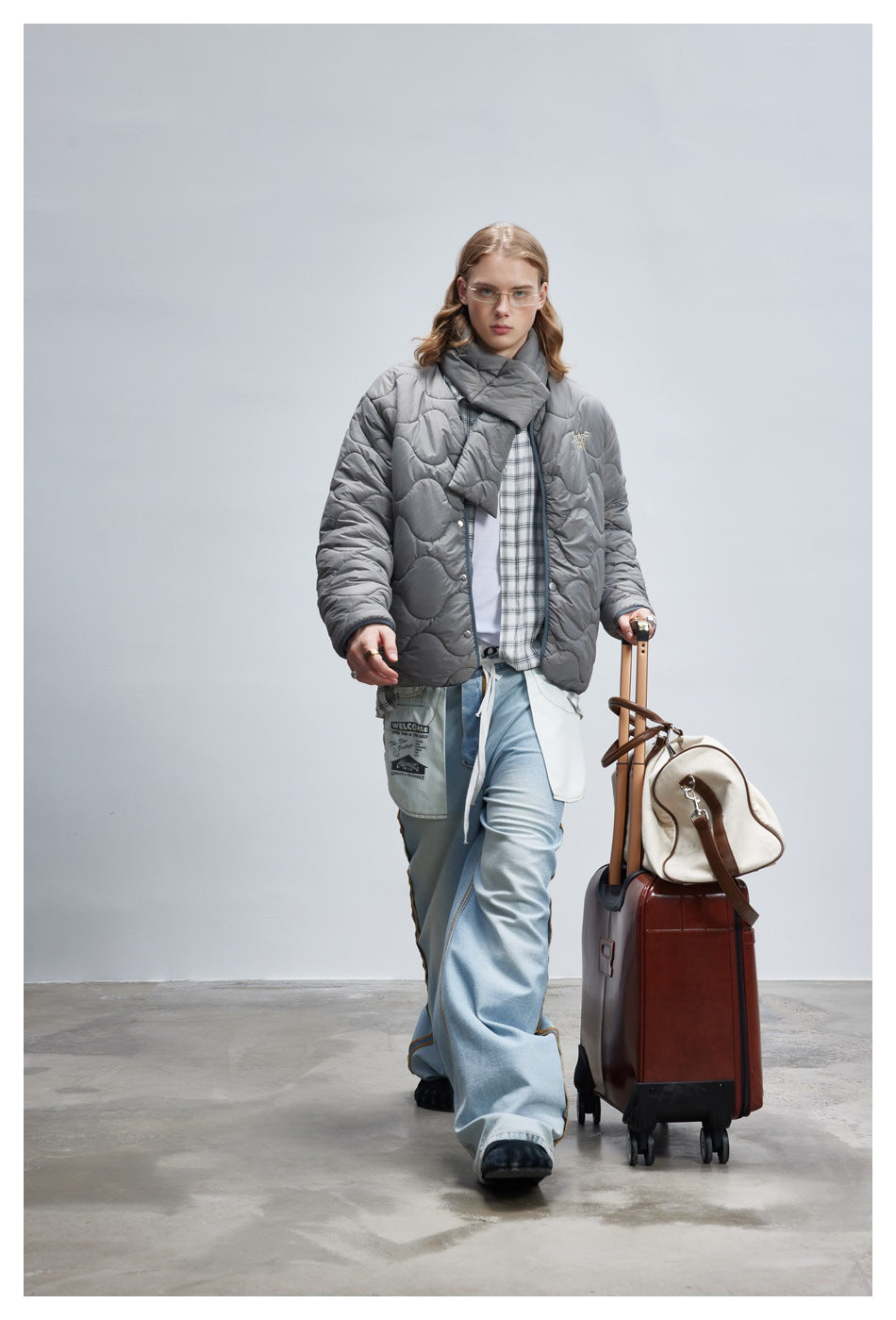 Model walking in a gray wave-quilted jacket and scarf, plaid shirt, and wide light-blue jeans.