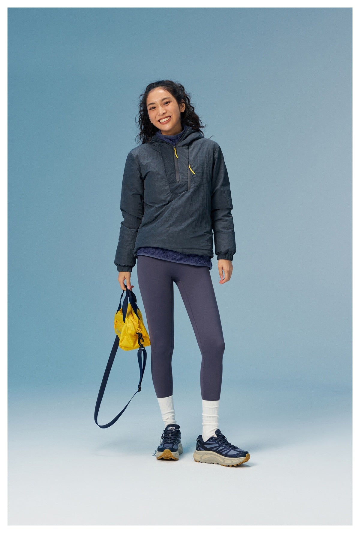 Woman wearing a charcoal hooded pullover with yellow zipper pulls and purple leggings, holding a yellow bag.