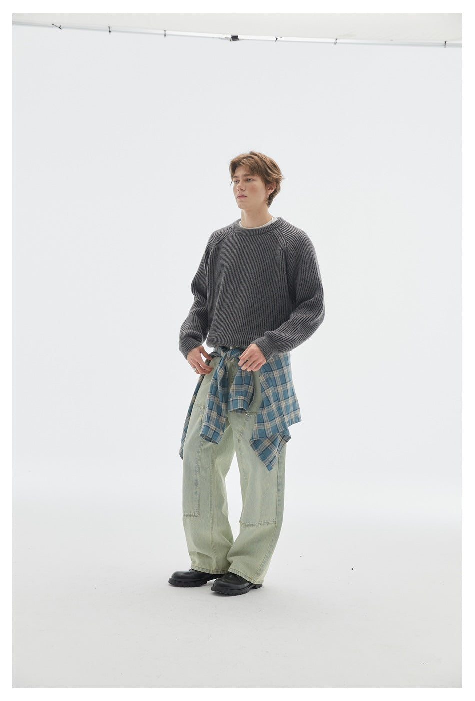 Three-quarter view of a model wearing a charcoal ribbed crewneck sweater with pale green jeans and a blue plaid shirt tied at